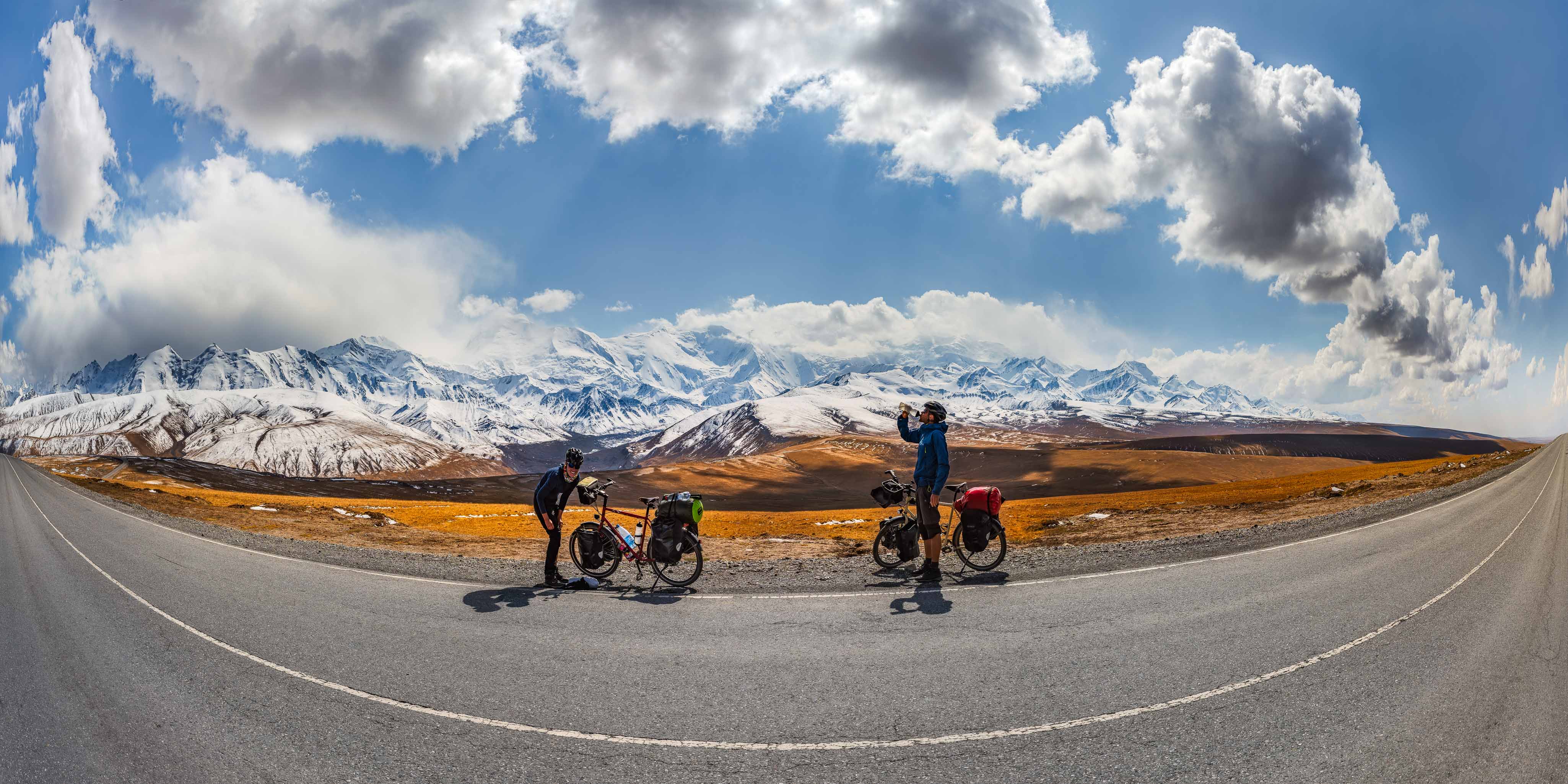 Zwei Fahrradfahrer verschnaufen vor der schneebedeckten Wand des Pamir in Kirgistan (Panorama).